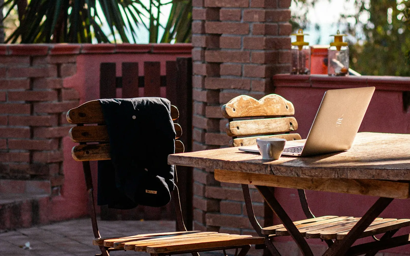An open laptop and a coffee cup on a weathered wooden table on a sunny brick terrace, with folding chairs, flowering shrubs and a palm behind